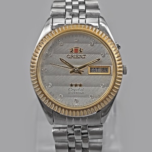 May include: Silver-toned Orient watch with a gold-colored bezel and a silver dial. The watch face features diamond-like markers and the day and date. The watch band is made of silver-colored metal links.