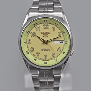May include: A silver-toned Seiko 5 automatic wristwatch with a light yellow dial. The watch features a day and date display, luminous hands and markers, and a stainless steel band. The watch face displays the text "SEIKO 5 AUTOMATIC 21 JEWELS".
