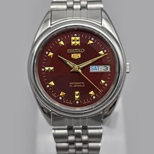 Könnte beinhalten: Eine silberfarbene Seiko 5 Automatik-Armbanduhr mit burgunderfarbenem Zifferblatt. Die Uhr hat goldfarbene Stundenmarkierungen und Zeiger, eine Tages- und Datumsanzeige und ein Edelstahlarmband. Das Zifferblatt zeigt "AUTOMATIC 21 JEWELS" und "MADE IN JAPAN".