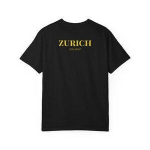 Könnte beinhalten: Schwarzes T-Shirt mit dem Wort "ZURICH" in goldenen Buchstaben auf dem oberen Rücken und dem Schriftzug "GITS EINE?" darunter. Das T-Shirt hat einen Rundhalsausschnitt und kurze Ärmel.