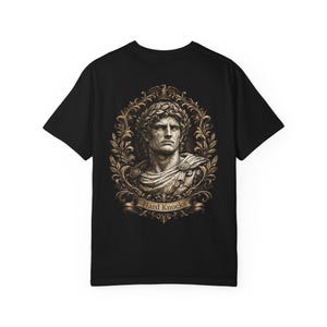 Könnte beinhalten: Ein schwarzes Kurzarm-T-Shirt mit einem Grafikdruck auf der Rückseite. Das Motiv zeigt eine steinerne Büste einer römisch anmutenden Figur mit Lorbeerkranz, eingefasst in einen verzierten goldfarbenen Rahmen mit dem Text Hard Knocks auf einem Banner.