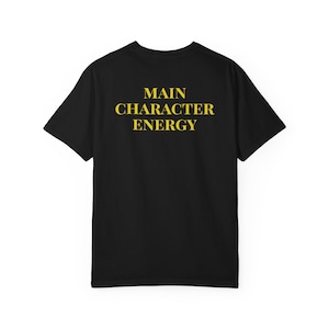 Könnte beinhalten: Schwarzes T-Shirt mit dem gelben Aufdruck "MAIN CHARACTER ENERGY". Der Text ist mittig auf der Rückseite des Shirts platziert. Das T-Shirt hat kurze Ärmel und einen Rundhalsausschnitt.