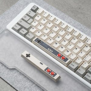 Peut inclure: Un clavier mécanique blanc avec des touches blanc cassé et des accents gris. La barre d'espace et un keycap séparé en forme de manette présentent un design de jeu vidéo rétro avec des boutons rouges. Le clavier repose sur une surface grise.