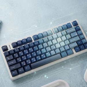 Puede incluir: Un teclado mecánico con marco blanco y teclas en degradado que pasan de azul marino oscuro a la izquierda a azul claro y blanco a la derecha. Las teclas muestran letras en inglés y caracteres coreanos Hangul en color blanco.