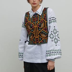 Puede incluir: Una camisa blanca de manga larga con mangas bordadas y un chaleco decorativo. El chaleco es negro con un diseño bordado con pájaros, hojas y ramas en tonos amarillos, naranjas y verdes.