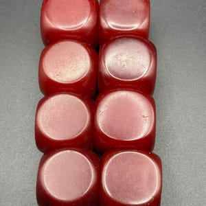 May include: Eight red dice arranged in two rows of four. Each die has rounded edges and a circular indentation on each face. The dice are a deep, translucent red colour, and are set against a dark grey background.