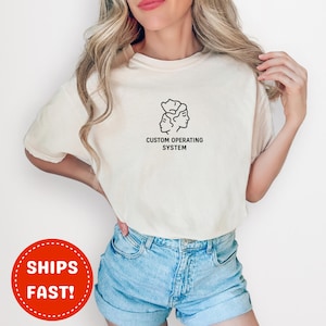 May include: Cream-colored t-shirt featuring a black line drawing of two faces and the text "CUSTOM OPERATING SYSTEM". The shirt is made of a soft material and has short sleeves.
