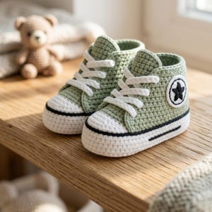 Può includere: Un paio di scarpine da neonato all'uncinetto, di colore verde chiaro. Le scarpe hanno lacci bianchi, una punta bianca e una striscia nera lungo la suola. Una stella nera è ricamata sul lato di ogni scarpa. Un orsacchiotto è sullo sfondo.