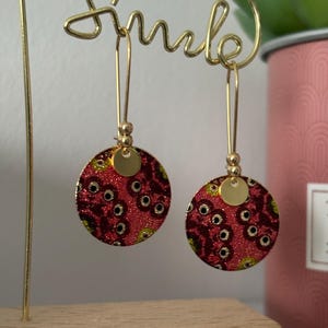 Puede incluir: Un par de pendientes colgantes dorados. Cada pendiente presenta un colgante circular con estampado rojo y dorado con detalles negros. Los pendientes se exhiben en un soporte de alambre dorado con la palabra "Smile" en cursiva.