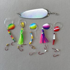 May include: A collection of fishing lures, including a large silver spoon lure and several smaller lures with colorful blades, beads, and hooks. The lures are designed for attracting fish.