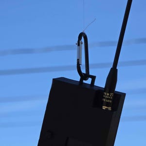 May include: A black rectangular device with a carabiner clip and antenna, suspended against a blue sky. The device has the text "mzi WORKS 915 MHz" printed on it.