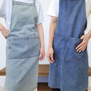 May include: Two denim aprons featuring a square neckline, thick shoulder straps, and a large front pocket with visible stitching. One apron is a light-wash blue denim, while the other is a medium-wash blue denim, both worn over white t-shirts.