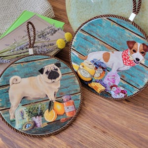May include: Two round hanging wall plaques featuring rustic blue wood backgrounds. One plaque displays a fawn pug with an orange, a jar, and a plant. The other plaque shows a Jack Russell terrier wearing a red polka dot scarf with a jar labelled Miel.