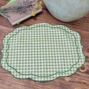 May include: A scalloped placemat featuring a green and white gingham check pattern. The fabric has a distinct scalloped edge finished with a thin green border, creating a soft, rounded rectangular shape that provides a classic textile appearance.