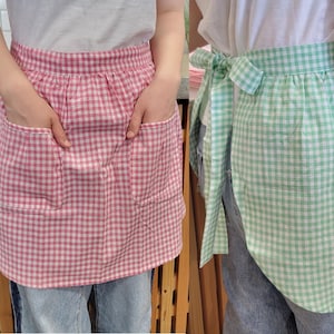 May include: Two half aprons in a gingham pattern. One is pink and white, the other is green and white. The aprons are worn over blue jeans. One apron is tied at the waist.