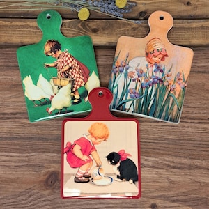 May include: Three rectangular ceramic chopping boards with rounded handles. One green board shows a child feeding chickens. One peach board features a child among purple irises. One red board depicts a child pouring milk for a black and white cat.