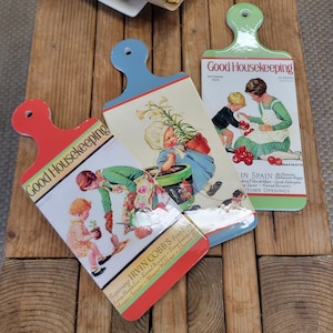 Puede incluir: Tres tablas de cortar vintage con temática de portadas de la revista Good Housekeeping. Cada tabla tiene un asa de diferente color: rojo, azul y verde. Las portadas presentan ilustraciones de niños y adultos, con texto como "October 1939" e "In Spain".