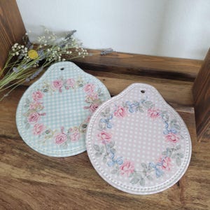 Könnte beinhalten: Zwei dekorative Keramik-Topfuntersetzer. Einer ist hellblau mit einem Vichy-Muster, der andere rosa mit einem gepunkteten Muster. Beide haben ein florales Rosenkranz-Design und ein Loch zum Aufhängen. Die Untersetzer liegen auf einer Holzoberfläche.