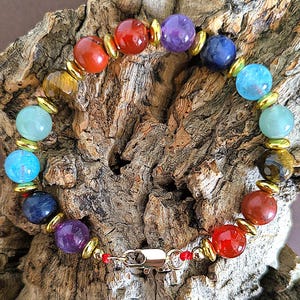 Puede incluir: Una pulsera de cuentas con una variedad de piedras preciosas redondas y pulidas en tonos rojo, naranja, amarillo, verde, azul y morado, intercaladas con pequeños espaciadores metálicos de color dorado y un cierre de mosquetón dorado.