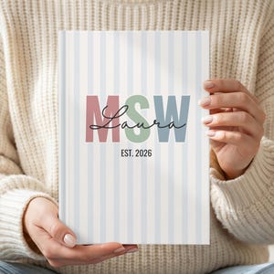 May include: A hardcover notebook with a light blue and white vertical striped cover. The center features large block letters M, S, and W in muted red, green, and blue, overlaid with the black cursive name Laura and the text EST. 2026.