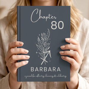 May include: A dark grey hardcover journal featuring the text "Chapter 80" at the top, a white line-art illustration of holly and a Sagittarius symbol in the centre, the name "BARBARA" below, and "a journal for reflecting, dreaming & celebrating".