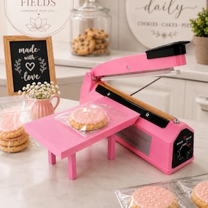 May include: A pink heat sealer with a cookie on a pink platform. The image also shows cookies in clear packaging, a chalkboard sign that says "made with love", and a sign that says "fresh baked daily".