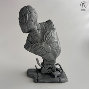 Puede incluir: Un busto gris de Spider-Man, con texturas detalladas de su traje y máscara. La escultura incluye un emblema de araña en el pecho y está montada sobre una base texturizada. Es un artículo de colección.