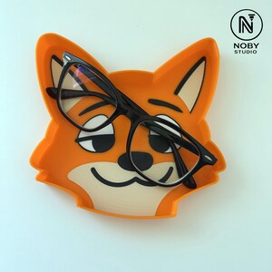 Peut inclure: Un plateau orange en forme de renard avec un visage souriant, tenant une paire de lunettes à monture noire. Le plateau a un intérieur beige clair et des détails noirs. Un objet amusant et fonctionnel pour organiser les lunettes.