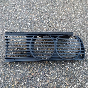 May include: A black plastic car air vent with horizontal slats and two circular vents. The vent has a rectangular shape with a foam strip attached to the top. The object is lying on a textured gray surface.