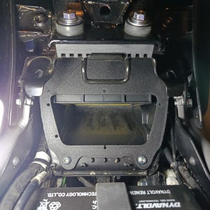 May include: An overhead view of a motorcycle's internal components. A black plastic airbox is visible, along with a battery and various wires and metal brackets. The battery has the text "DYNAVOLT RENEW" and "EXPE" printed on it.