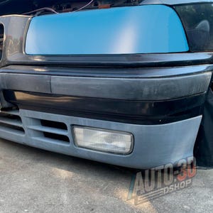 May include: Close-up of a car's front end, featuring a blue film over the headlights and a gray bumper. The bumper has a fog light and a lower air intake. The car is black, with the text "AUTO-3D EDSHOP" visible.