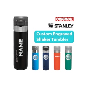 May include: A black Stanley shaker tumbler with a silver lid and the name "NAME" printed vertically in white. The image also shows four other shaker tumblers in blue, navy, orange, and green, each with custom text.