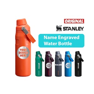 Peut inclure: Une gourde Stanley orange avec une poignée argentée et un autocollant rond avec l'inscription "YOUR DESIGN HERE". L'image montre également cinq autres gourdes en vert, violet, gris, bleu et noir. Le texte "Name Engraved Water Bottle" est affiché.