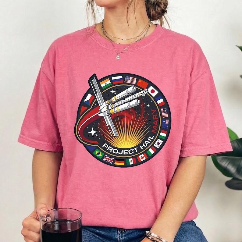 Pu&ograve; includere: T-shirt rosa con una grafica circolare raffigurante una navicella spaziale, bandiere e la scritta "PROJECT HAIL MARY". Il design ha un tema spaziale, con un sole giallo e uno sfondo stellato.