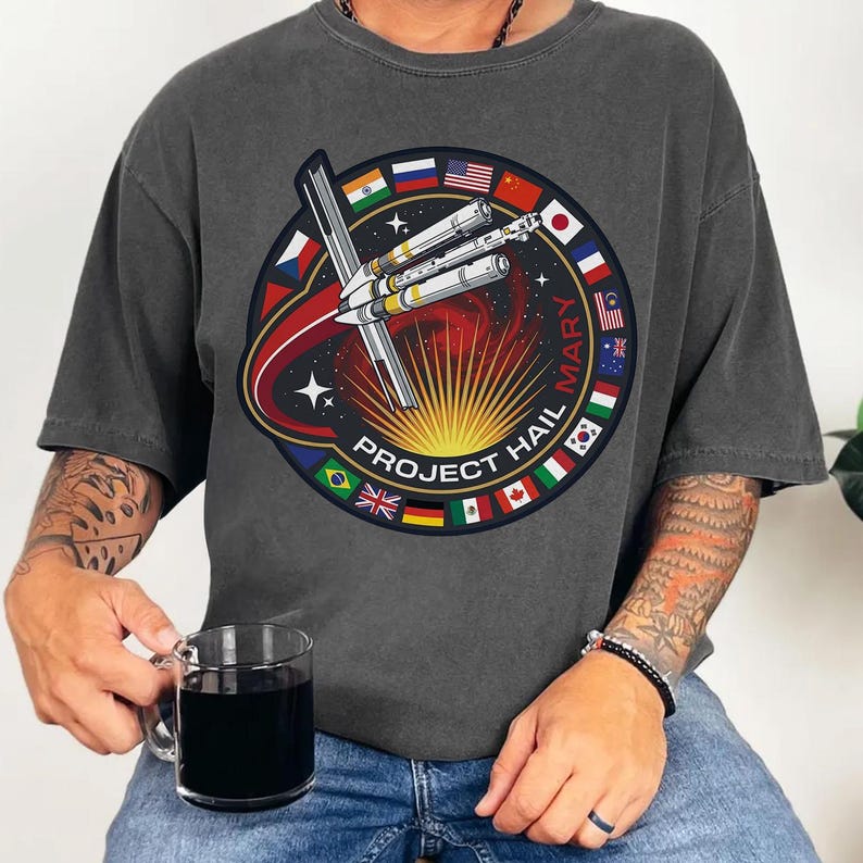 Pu&ograve; includere: T-shirt grigio scuro con una grafica circolare raffigurante un razzo e la scritta "PROJECT HAIL MARY". Il design incorpora bandiere di diversi paesi. Una persona che indossa la maglietta tiene in mano una tazza di vetro trasparente.
