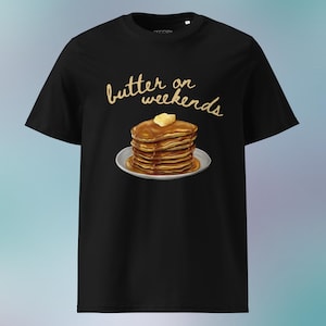 Könnte beinhalten: Schwarzes T-Shirt mit einem Pfannkuchen-Motiv auf einem weißen Teller, beträufelt mit Sirup und mit Butter belegt. Der Text "butter on weekends" steht in goldener Schrift über den Pfannkuchen. Ein lässiges, Food-Themen-Grafik-T-Shirt.