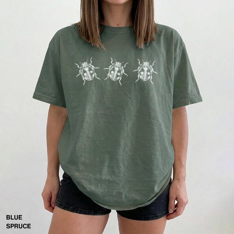 May include: A sage green short-sleeve t-shirt featuring a white graphic print of three ladybugs arranged in a horizontal row across the chest. The shirt has a crew neckline and a relaxed fit, with the color labeled as Blue Spruce.