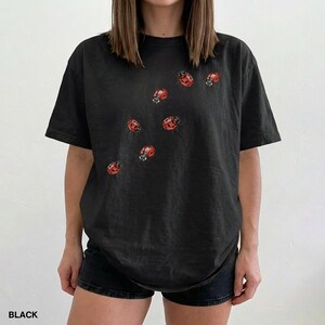 Może przedstawiać: Czarny t-shirt z wzorem czerwonych biedronek. Biedronki są rozrzucone po przodzie koszulki. Słowo "BLACK" jest wydrukowane na dole obrazu. Koszulka ma kr&oacute;tkie rękawy i luźny kr&oacute;j.