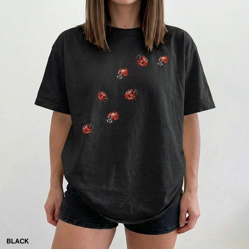 Może przedstawiać: Czarny t-shirt z wzorem czerwonych biedronek. Biedronki są rozrzucone po przodzie koszulki. Słowo "BLACK" jest wydrukowane na dole obrazu. Koszulka ma kr&oacute;tkie rękawy i luźny kr&oacute;j.
