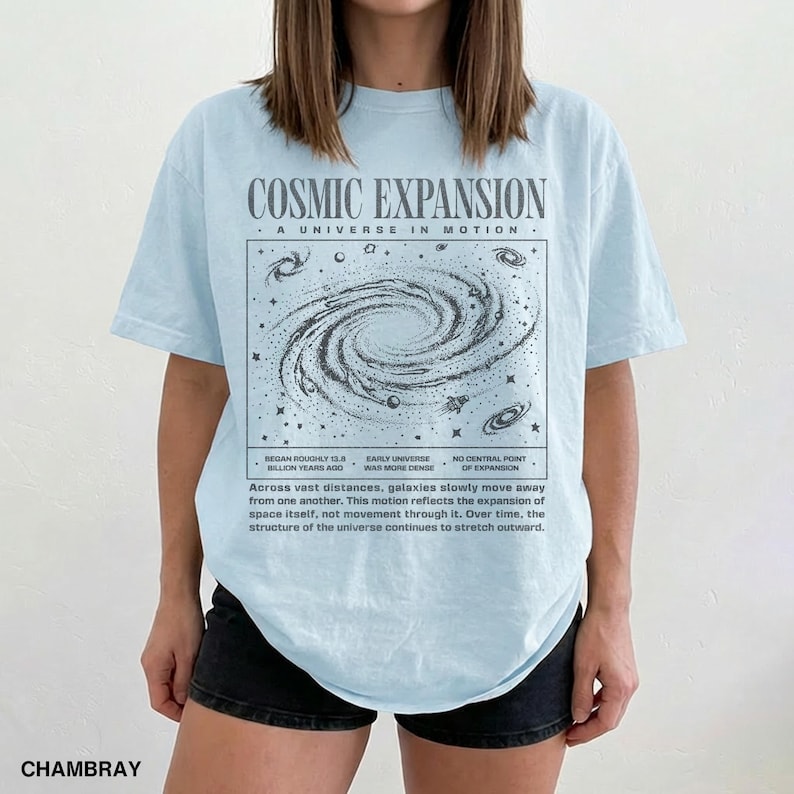 May include: Light blue t-shirt with a graphic of a galaxy and the text "COSMIC EXPANSION". The shirt also includes text about the universe's expansion. The shirt is made of chambray fabric.