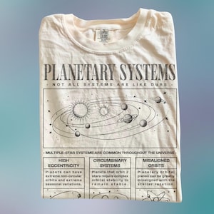 May include: Cream-colored t-shirt with a graphic of a planetary system. The design includes the text "PLANETARY SYSTEMS" and scientific diagrams. The shirt also features the text "NOT ALL SYSTEMS ARE LIKE OURS" and information about different types of planetary systems.