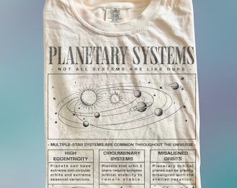 May include: Cream-coloured t-shirt featuring a planetary system graphic. The design includes the text "PLANETARY SYSTEMS" and scientific diagrams. The shirt also features the text "NOT ALL SYSTEMS ARE LIKE OURS" and information about planetary systems.