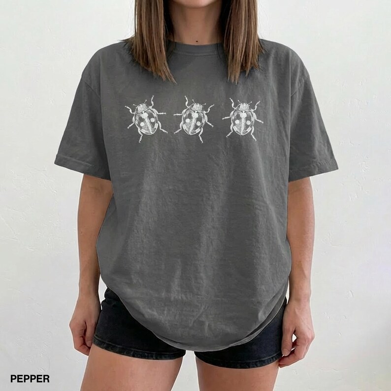 May include: A charcoal gray short-sleeve t-shirt featuring a horizontal row of three white line-art illustrations of ladybugs centered on the chest. The shirt has a crew neckline and a relaxed fit, constructed from a solid-colored cotton fabric.