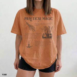 May include: An orange t-shirt with the words "PRACTICAL MAGIC" printed across the chest. The shirt features a detailed graphic of herbs, bottles, and a candle. The shirt is made of a soft material and is a relaxed fit.
