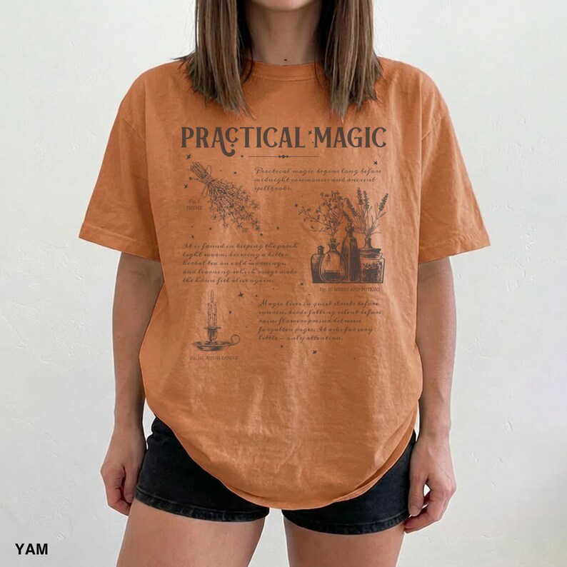 May include: An orange t-shirt with the words "PRACTICAL MAGIC" printed across the chest. The shirt features a detailed graphic of herbs, bottles, and a candle. The shirt is made of a soft material and is a relaxed fit.