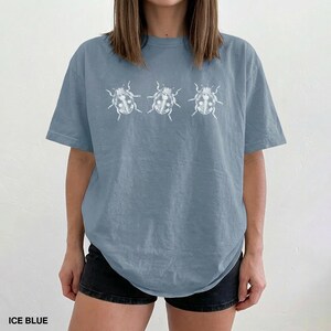 May include: An ice blue short-sleeved t-shirt featuring a horizontal row of three white line-art illustrations of beetles printed across the chest. The garment has a crew neckline and a relaxed fit, showcasing a simple graphic design.