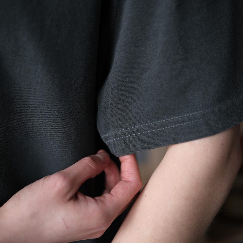 May include: A dark charcoal gray t-shirt featuring short sleeves with visible double-stitched hems. The fabric has a matte, slightly textured appearance, and the garment is constructed with a standard crew neckline and a relaxed, loose-fitting silhouette.
