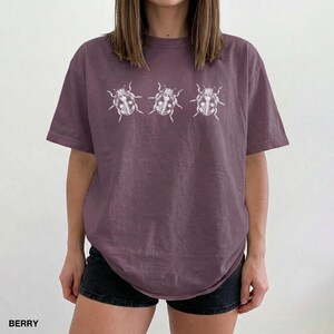 May include: A berry-colored short-sleeve t-shirt featuring a white graphic print of three ladybugs arranged in a horizontal row across the chest. The shirt has a crew neckline and a relaxed fit, with the word BERRY printed in the bottom corner.