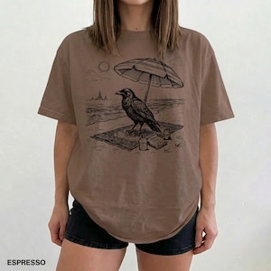 May include: Espresso-colored t-shirt featuring a black graphic of a raven relaxing on a beach under a striped umbrella. The design includes a beach scene with a sun, waves, and a small bottle. The word "ESPRESSO" is printed at the bottom.