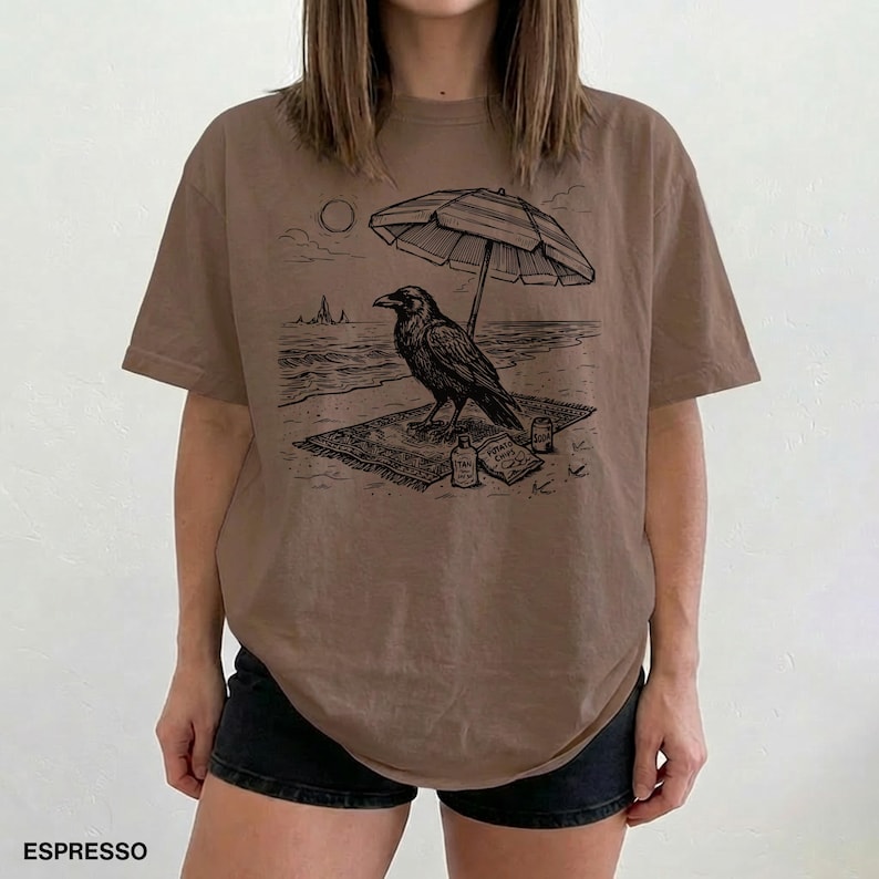 May include: Espresso-colored t-shirt featuring a black graphic of a raven relaxing on a beach under a striped umbrella. The design includes a beach scene with a sun, waves, and a small bottle. The word "ESPRESSO" is printed at the bottom.